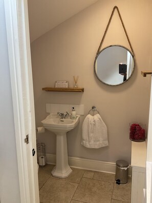 Un baño con lavamanos pedestal, un espejo, barra para toallas y un cubo de basura.