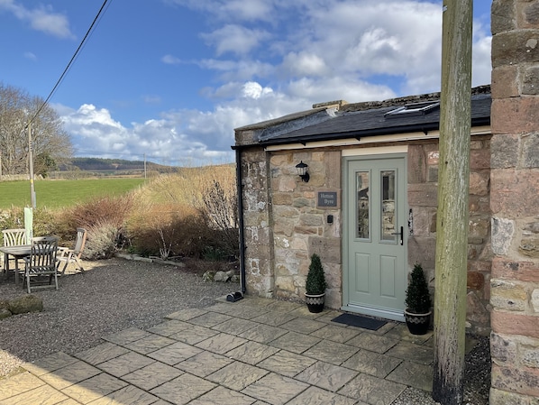 A stone cottage with a green door, a small outdoor seating area, and a view of the countryside.