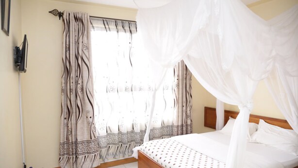 A bedroom with a four-poster bed, a curtain with a floral pattern, and a bedside table with a remote control and water bottles. - Roza Guests House (Naivasha)