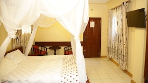 A room with a bed, a TV, a chair, a table, and curtains. - Roza Guests House (Naivasha)