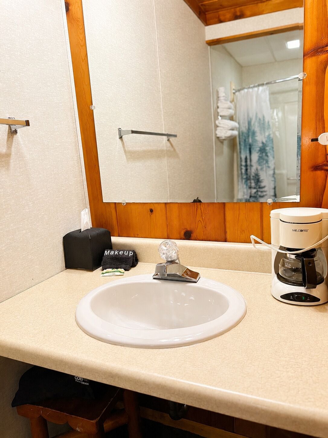 Een badkamer met een witte wastafel, een koffiezetapparaat, een toilettas en een spiegel die een douchegordijn weerkaatst.