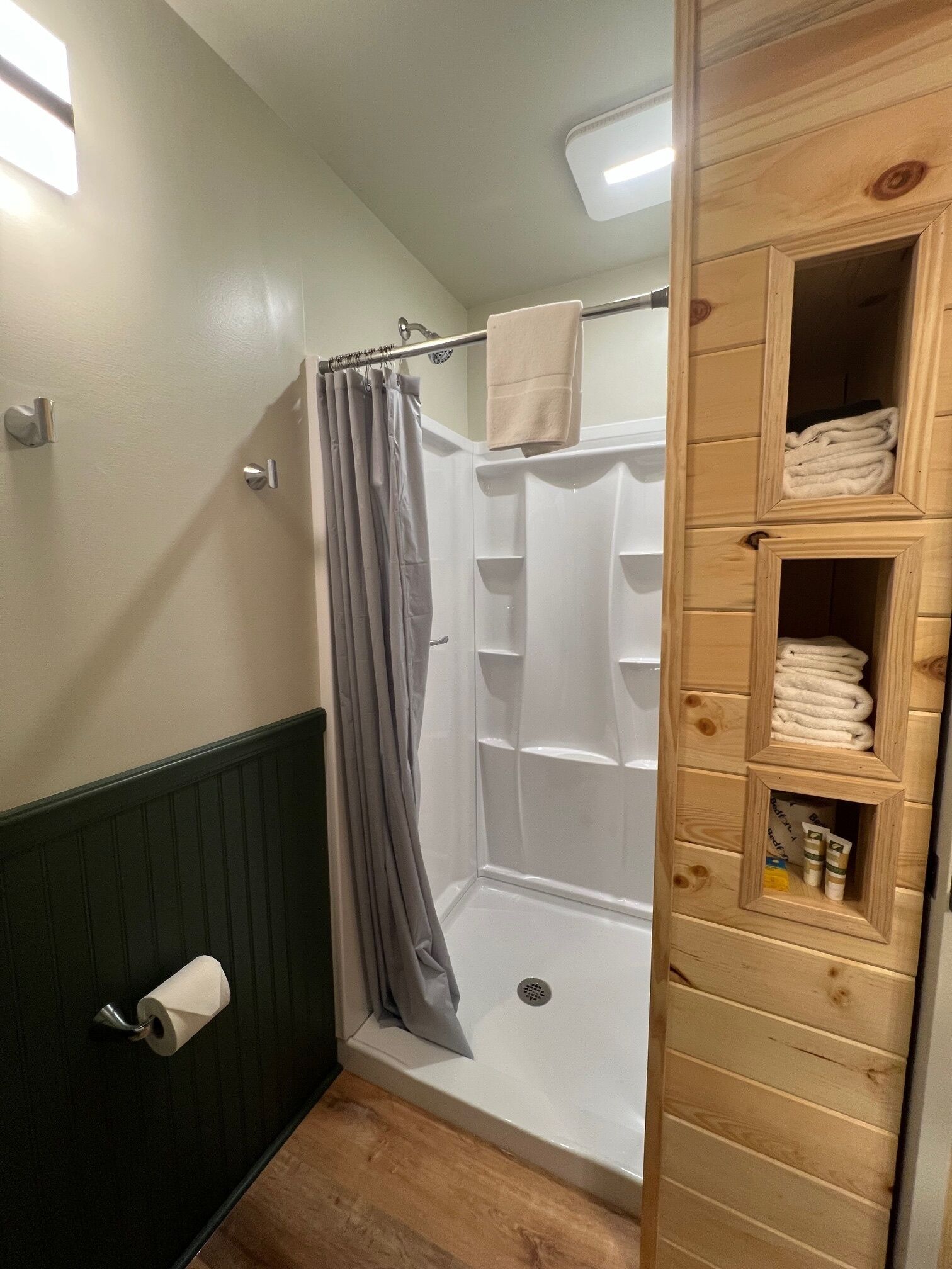 A bathroom with a shower, a towel rack, a toilet paper holder, and a wooden shelf with towels and toiletries.