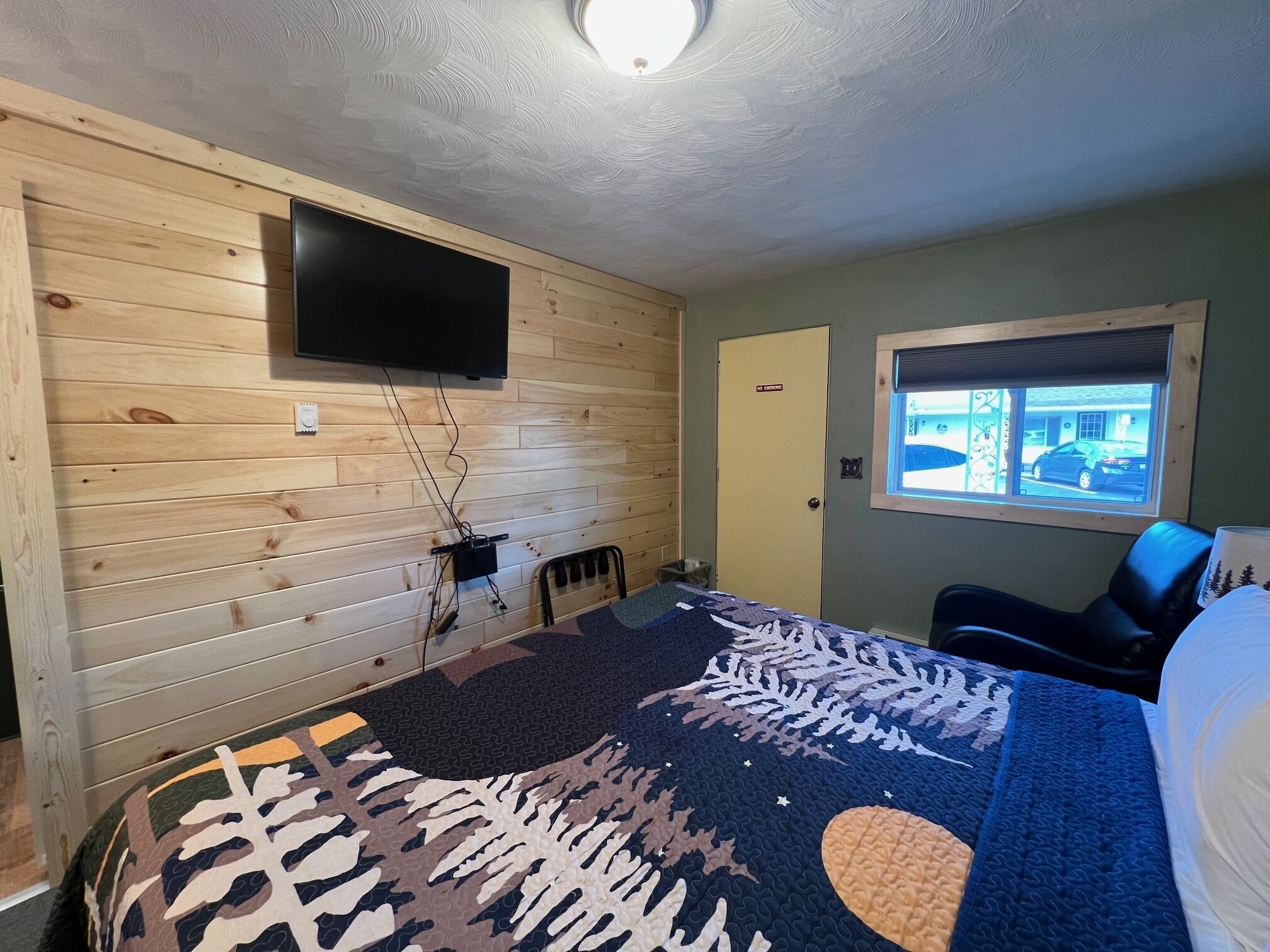 A room with a bed, a television mounted on the wall, a window with a view of a car outside, and a door with a 'No Smoking' sign.