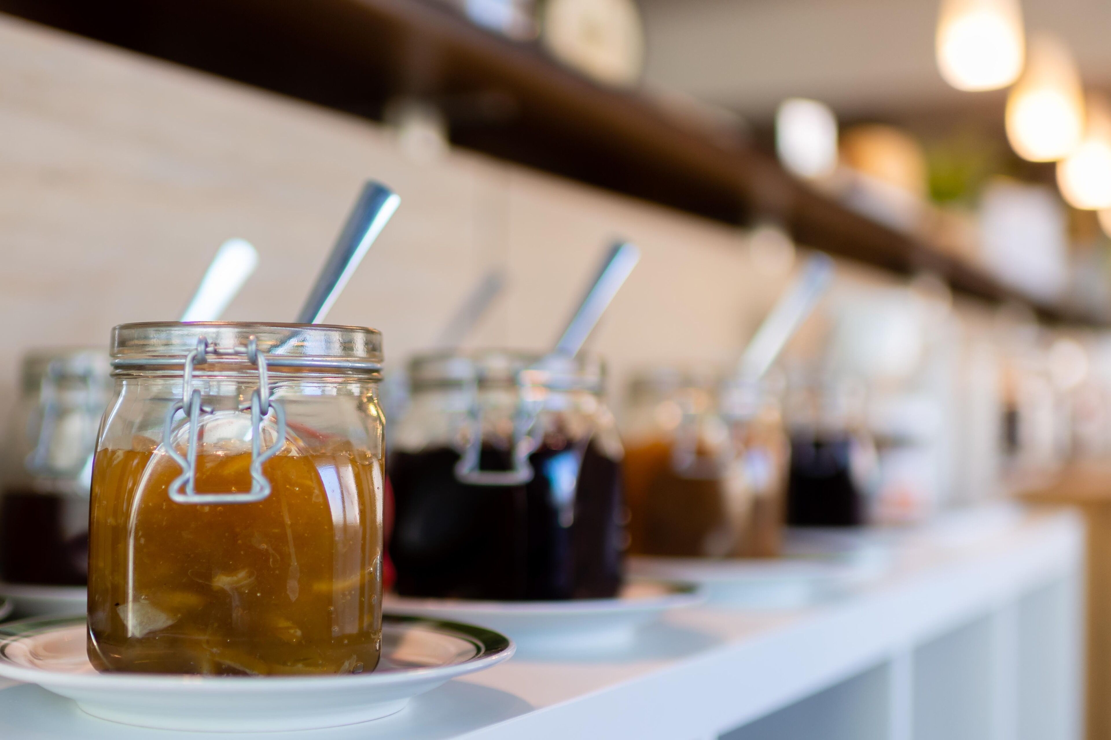 A row of glass jars with metal lids and spoons, likely containing beverages, on a white surface.