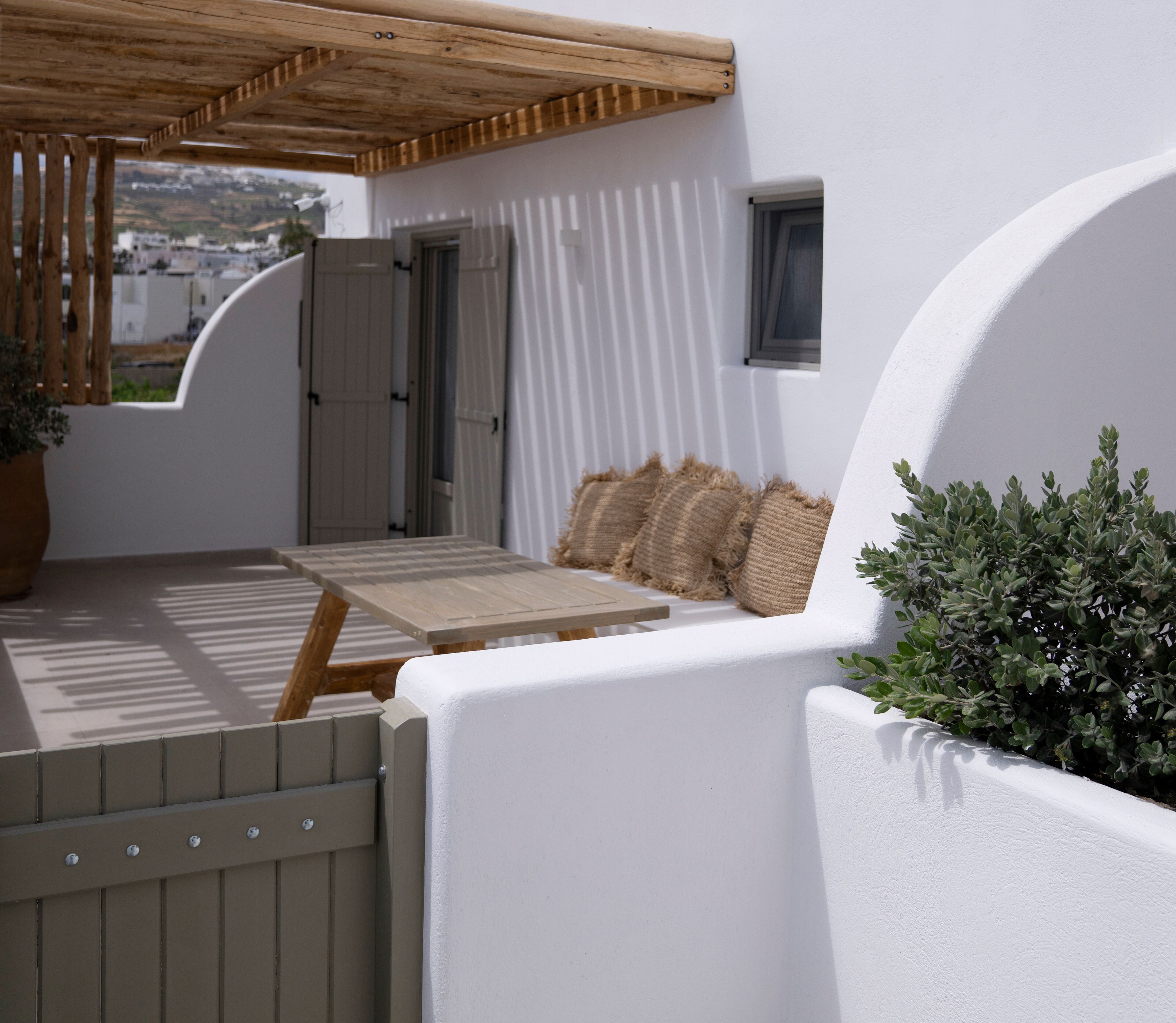 Un espace extérieur aménagé avec des murs blancs, comprenant une table en bois et des coussins, une pergola en bois et un pot de fleurs.