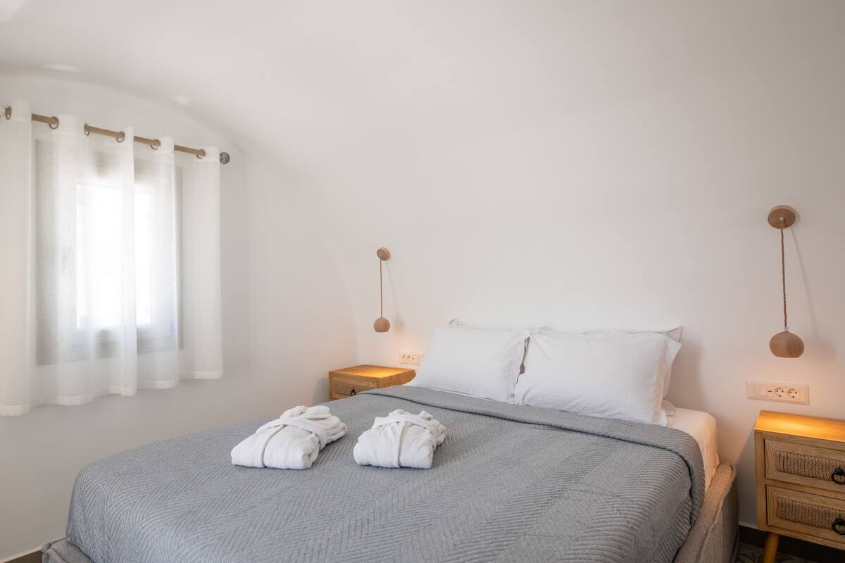 Una cama bien hecha con dos toallas, una mesita de noche con una lámpara y una ventana con cortinas translúcidas.