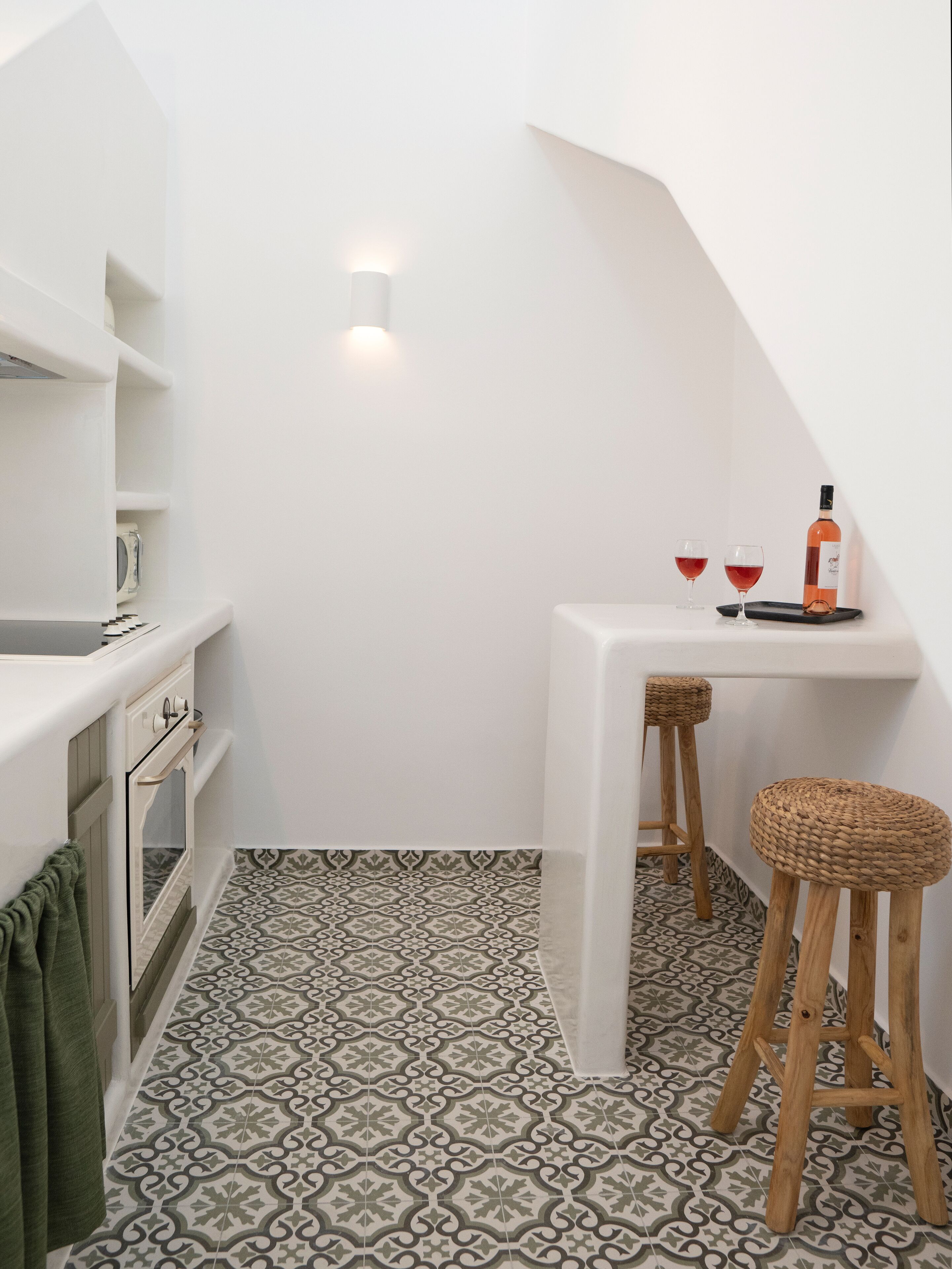 A compact kitchen with white cabinetry, a patterned floor, and a small dining area with stools and a wine bottle.
