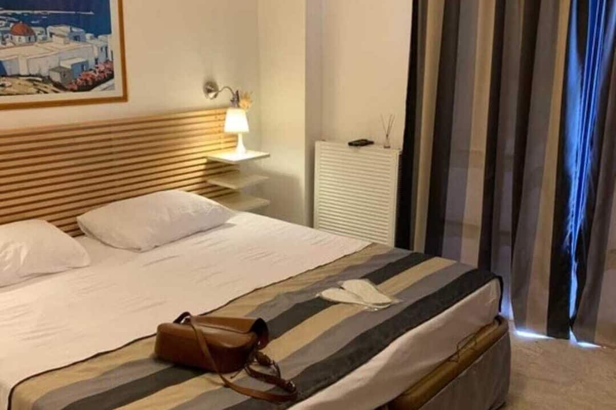 Quarto de hotel com uma cama, mesa de cabeceira com abajur, um quadro na parede e uma janela com cortinas.