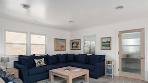 A living room with a dark blue sectional sofa, a wooden coffee table, and a patterned rug. There are framed pictures on the wall, a window with a view of a brick building, and a door with a frosted glass panel.