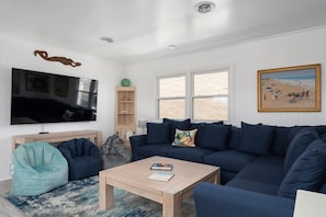 Uma sala de estar com um sofá azul escuro, uma mesa de centro de madeira, um assento de bola verde-água, uma TV, uma estante de livros e uma pintura de uma cena de praia.