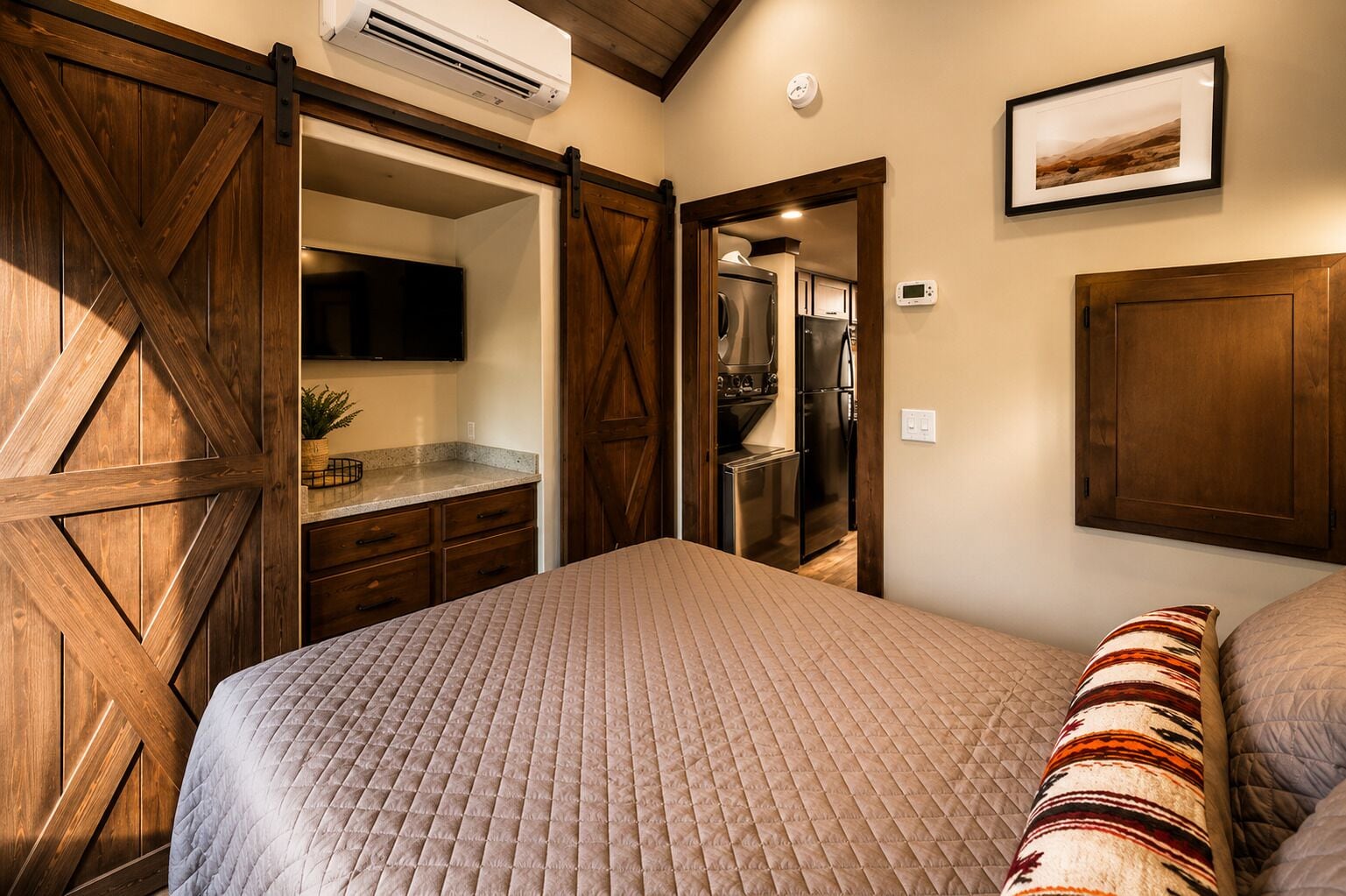 A bedroom with a large bed, a wardrobe, a built-in air conditioning unit, a kitchen area, and a framed picture on the wall.