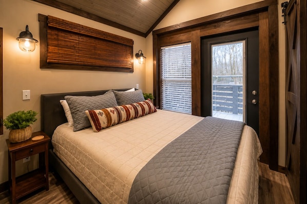A bedroom with a bed, bedside tables, a wall-mounted light fixture, and a door with a view of the outside.