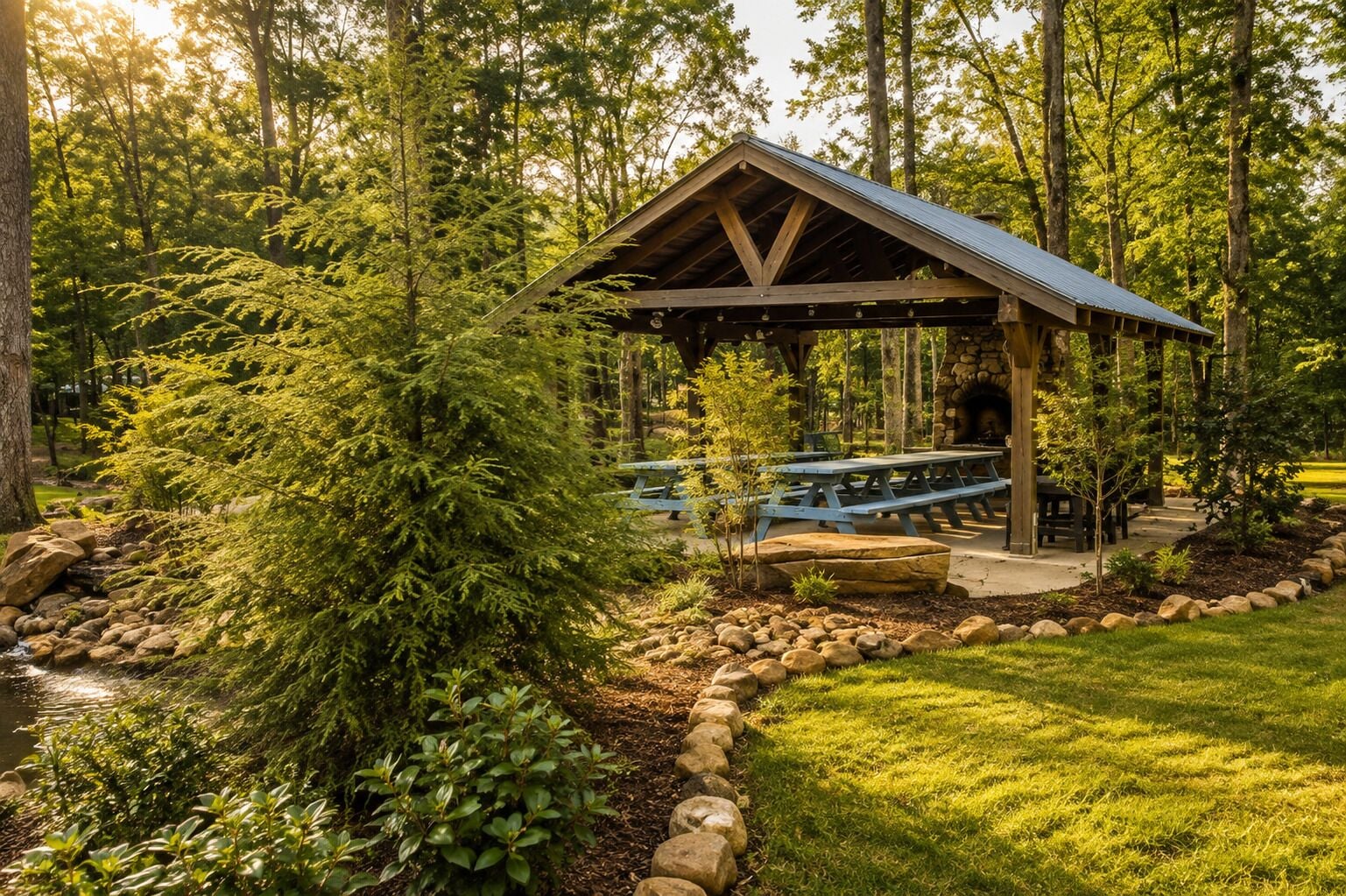 Un pavillon rustique en bois, doté de tables de pique-nique, est entouré d’un paysage naturel composé d’arbres et d’un petit ruisseau.