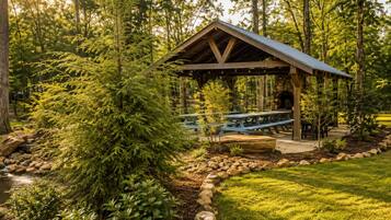Un pavillon rustique en bois, doté de tables de pique-nique, est entouré d’un paysage naturel composé d’arbres et d’un petit ruisseau.
