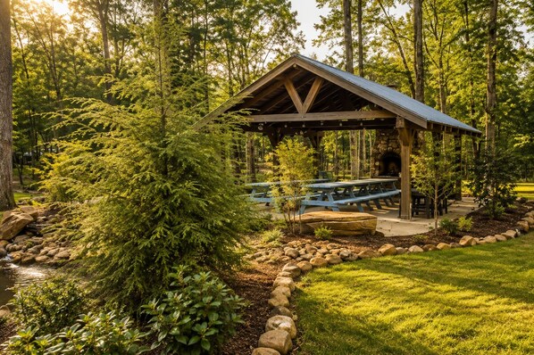 A rustic wooden pavilion with picnic tables, surrounded by a natural landscape with trees and a small stream.
