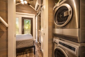 A compact laundry room with stacked washing machines and dryers, a bed with a quilted mattress, and a door leading to an outdoor area with a view of trees.