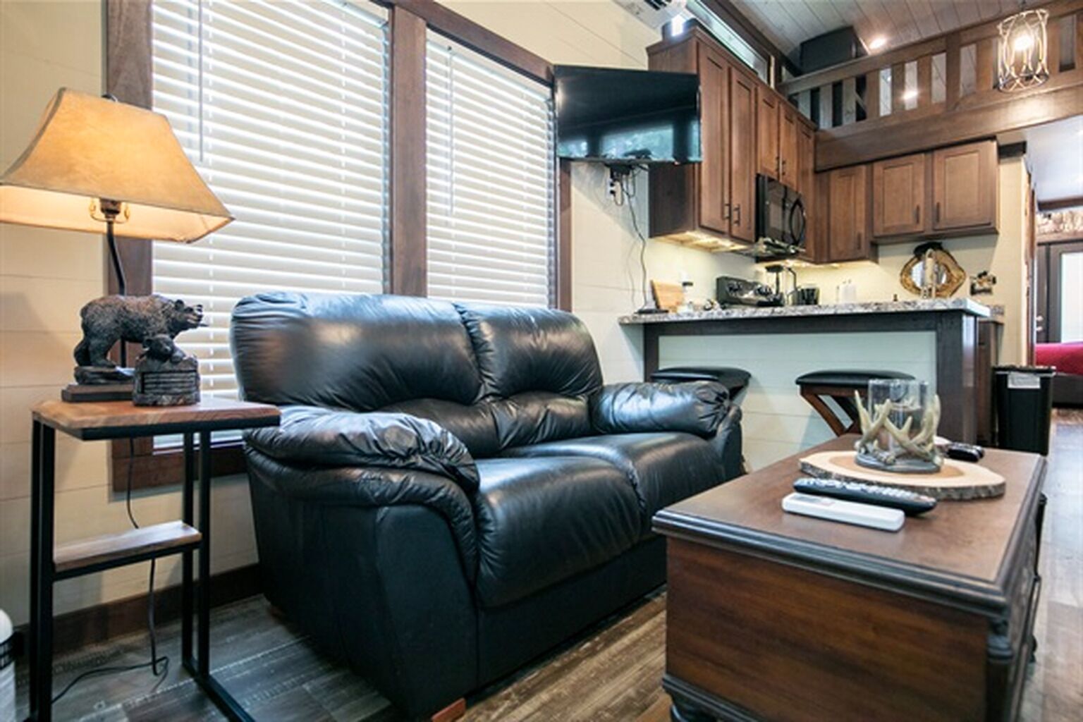 A living room with a black leather sofa, a small table with a lamp, and a kitchen area in the background.