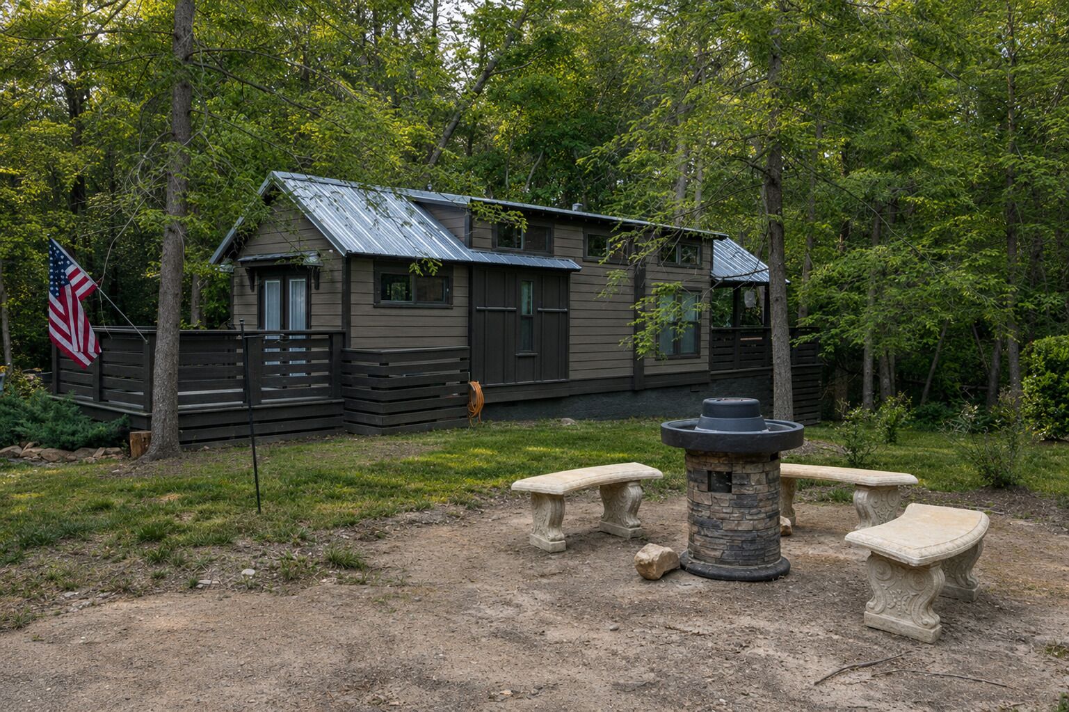 A cabin with a metal roof, surrounded by trees, and featuring a stone fire pit with benches.