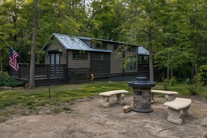 Una cabaña con techo de metal, rodeada de árboles, y con una chimenea de piedra con bancos.