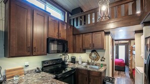 A kitchen with wooden cabinets, a black microwave, a stove, granite countertops, and a hanging light fixture. - Jenny's Retreat at The Retreat at Deer Lick Falls (Monteagle)