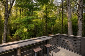 Une terrasse en bois avec une rambarde, deux tabourets et un banc donnant sur une forêt.
