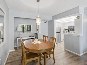 A dining area with a round wooden table, chairs, a chandelier, and a view into the kitchen. - Beachfront 2BR/2BA condo with pool, WiFi, and heated pool access. (Marco Island)