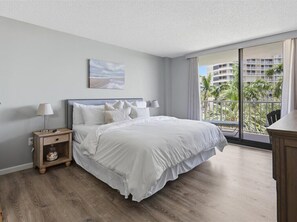 Une chambre avec un grand lit, une table de chevet, une lampe, un tableau au mur et un balcon offrant une vue sur les palmiers et les immeubles.
