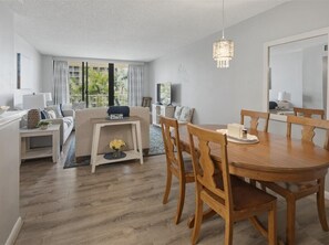 Una sala de estar moderna con comedor, que incluye una mesa y sillas de madera, un sofá, una mesa de centro y un amplio ventanal con cortinas.