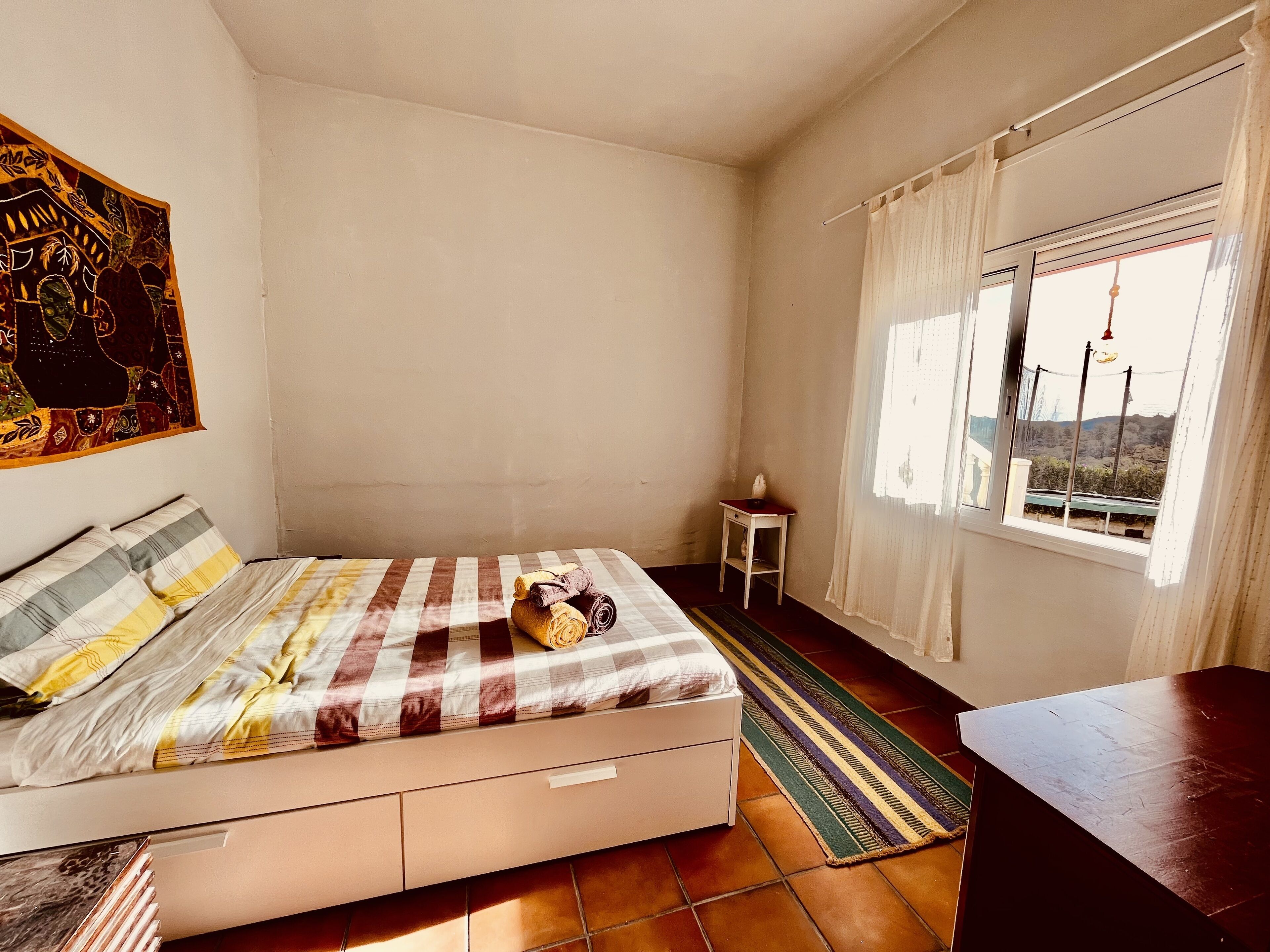 A bedroom with a bed, a small table, a window with curtains, and a view of the outside.