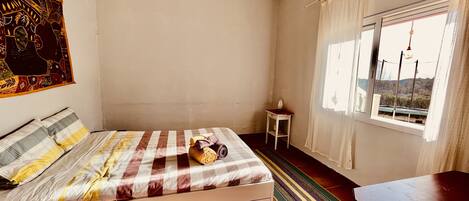 A bedroom with a bed, a small table, a window with curtains, and a view of the outside.