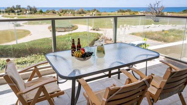 Een balkon met een glazen tafel en houten stoelen, met uitzicht op een aangelegde tuin en de oceaan.