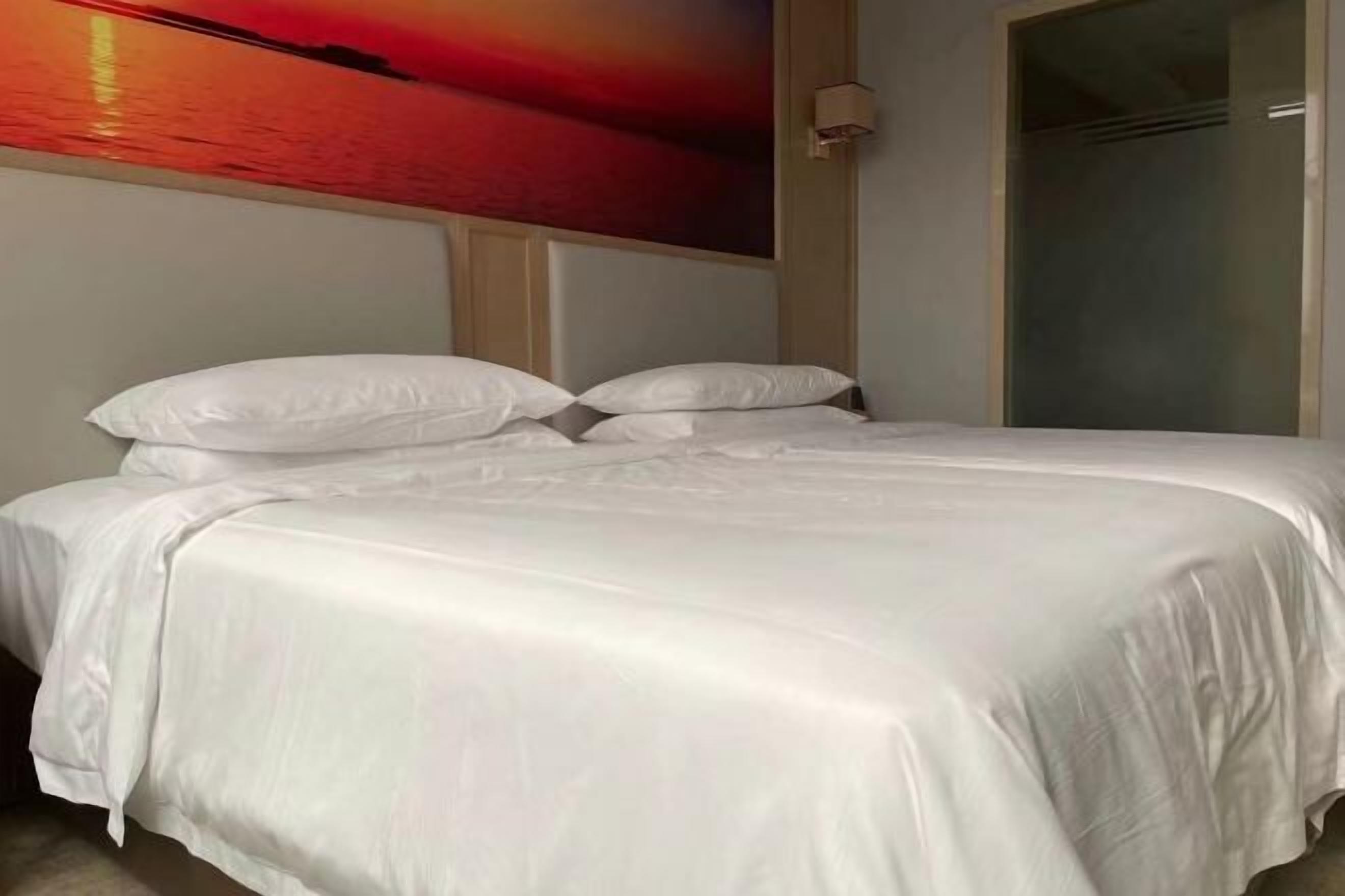 Un lit bien fait, avec du linge de lit blanc, dans une pièce où un grand tableau représentant un coucher de soleil sur l’eau est accroché au mur.