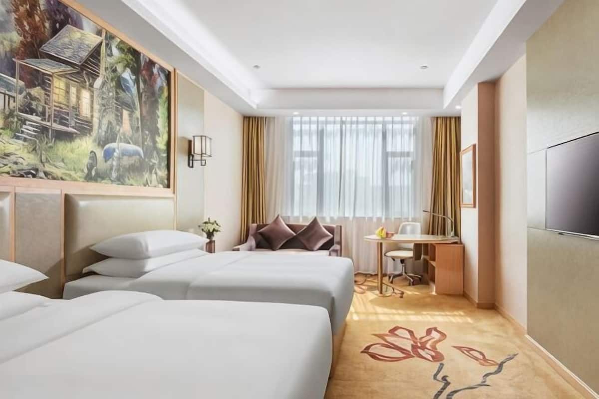 A hotel room with three beds, a desk, a chair, a TV, and a large painting on the wall.