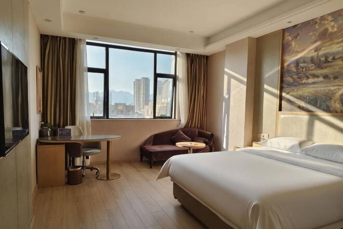 A hotel room with a bed, a desk, a chair, a small table, and a large window with a city view.