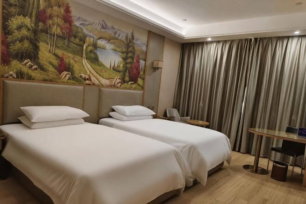 A hotel room with two beds, a desk, a chair, a painting, and a view of a mountain landscape.