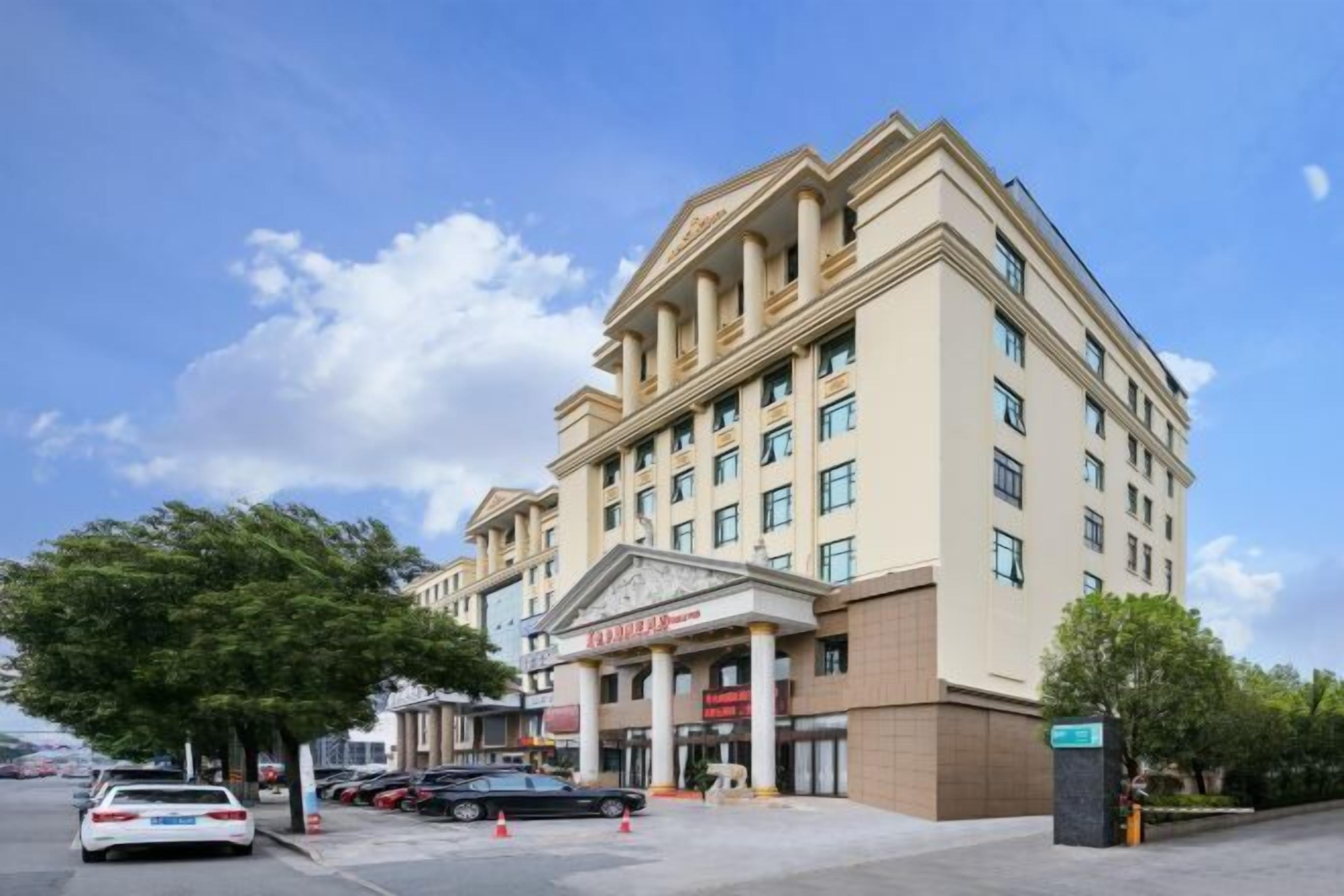 A multi-story hotel with a classical facade, featuring a central entrance with columns, multiple windows, and a parking area with cars.