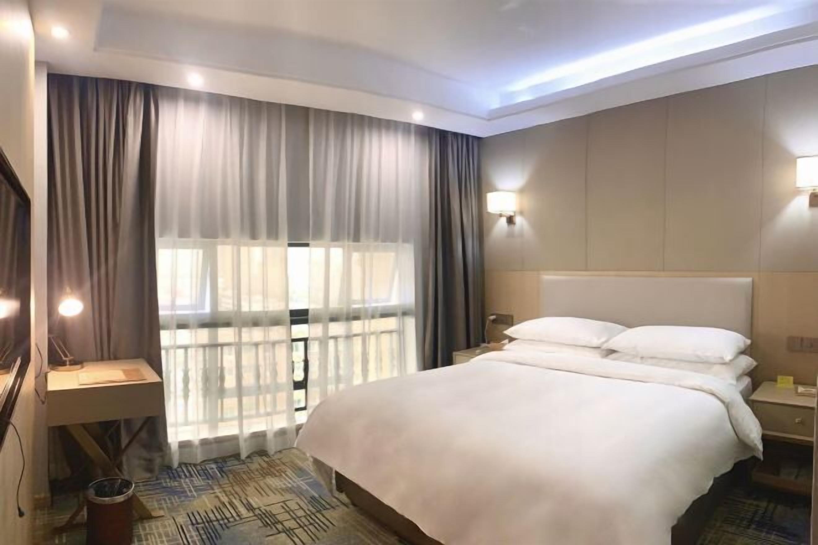 Une chambre d’hôtel avec un grand lit, un bureau avec une lampe, une fenêtre avec des rideaux et un luminaire fixé au mur.