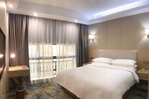 A hotel room with a large bed, a desk with a lamp, a window with curtains, and a wall-mounted light.