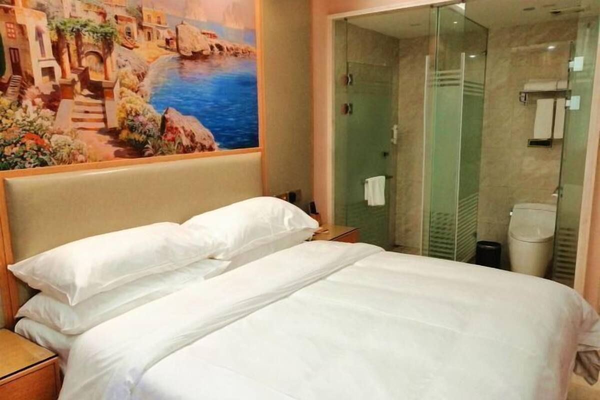 A hotel room with a bed, a painting of a coastal scene, and a glass-enclosed shower area.