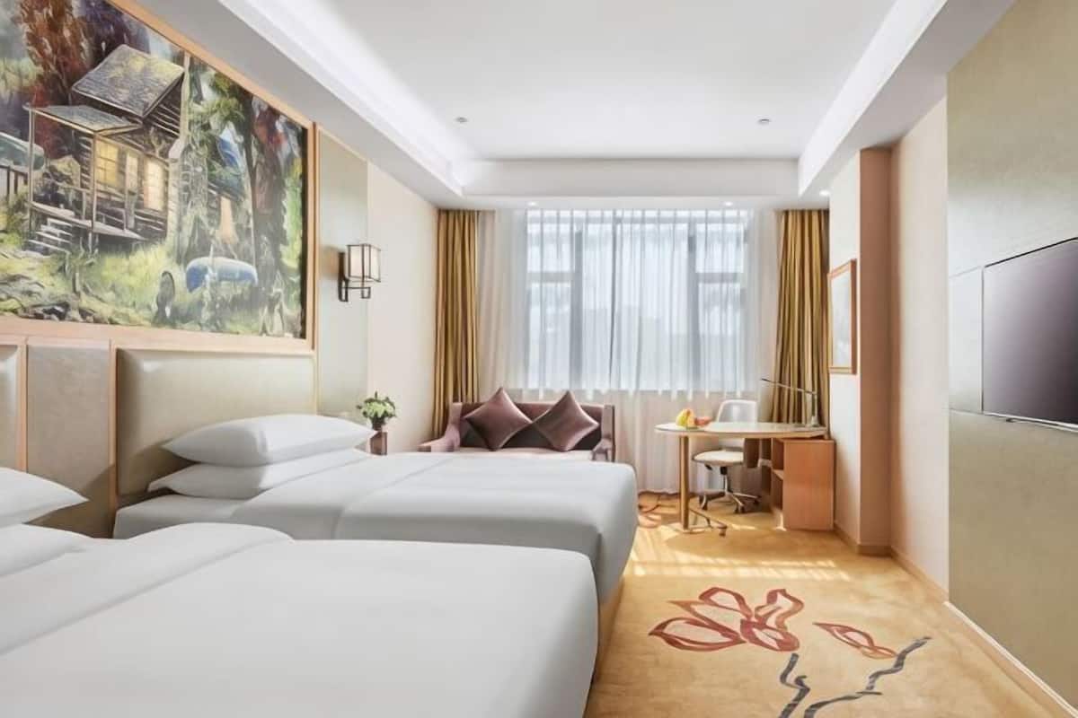 A hotel room with three beds, a desk, a chair, a TV, and a painting on the wall.