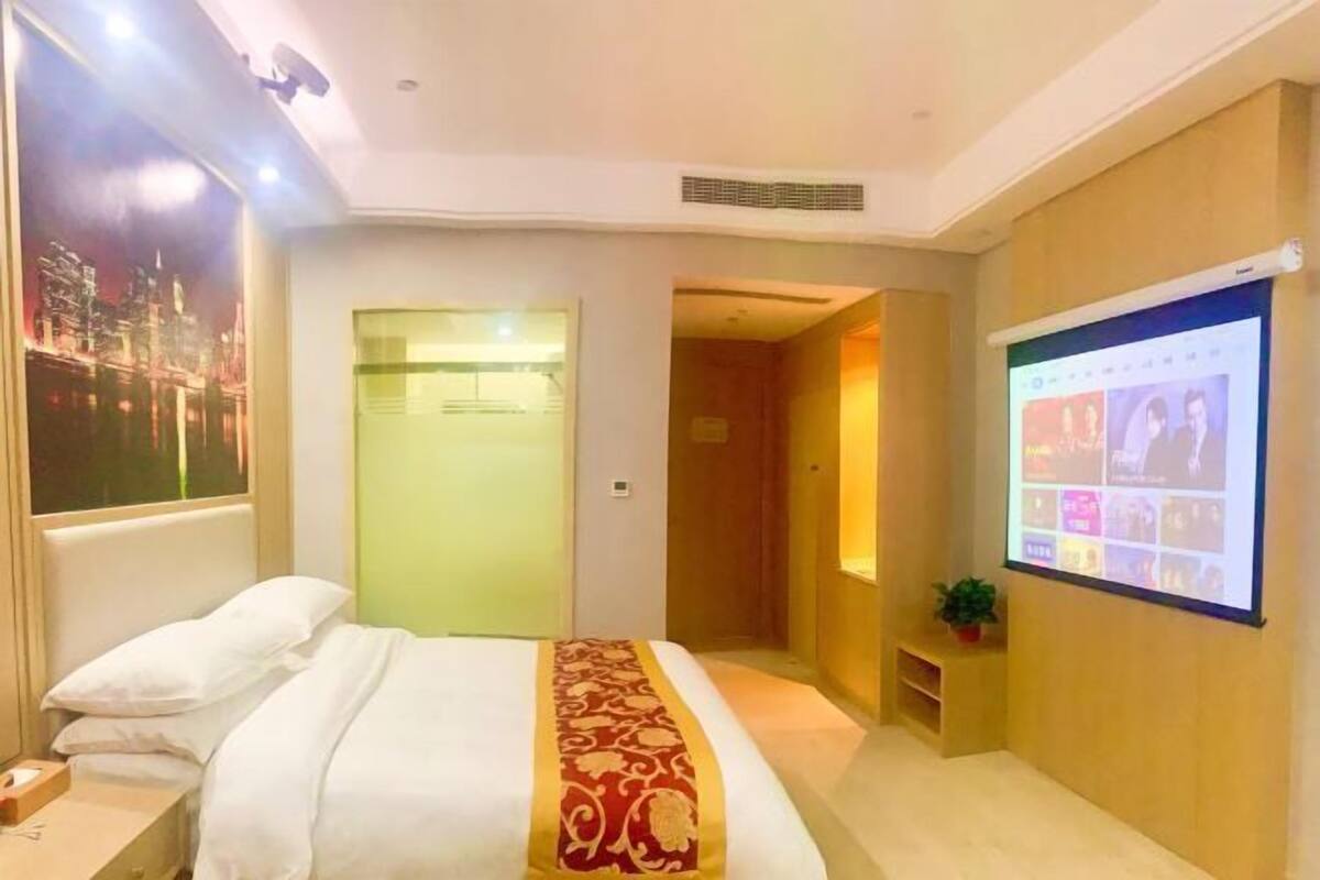 A hotel room with a bed, a projector screen, a small table with a plant, and a door leading to another room.