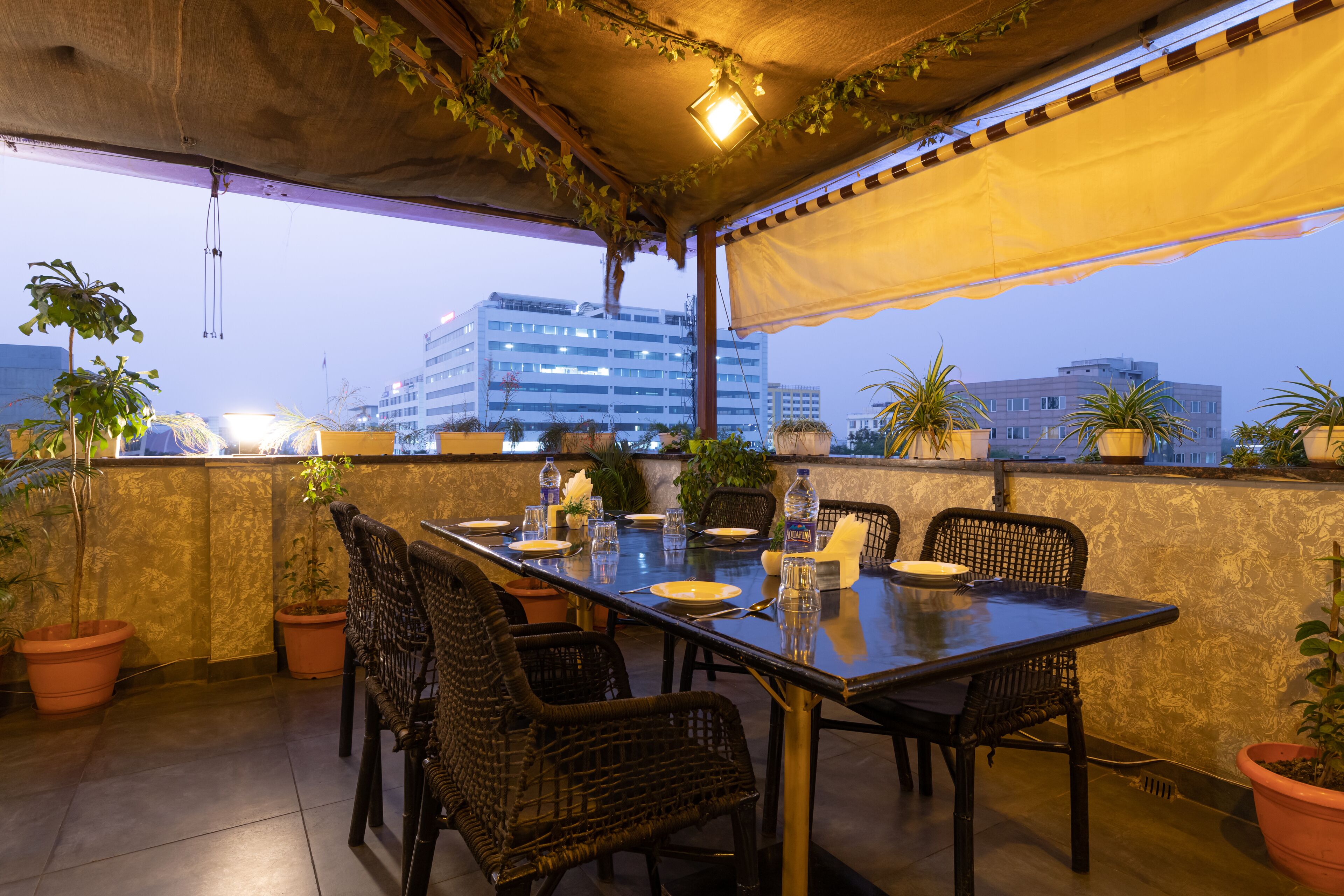 Un área de comedor al aire libre con una mesa preparada para cuatro personas, rodeada de sillas de mimbre, plantas en macetas y vistas de edificios al fondo.