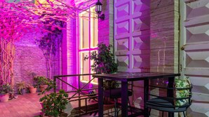 Une cour avec un sol à motifs, une table, des chaises, des plantes en pot et un bâtiment doté d’une façade en bois et d’une fenêtre.