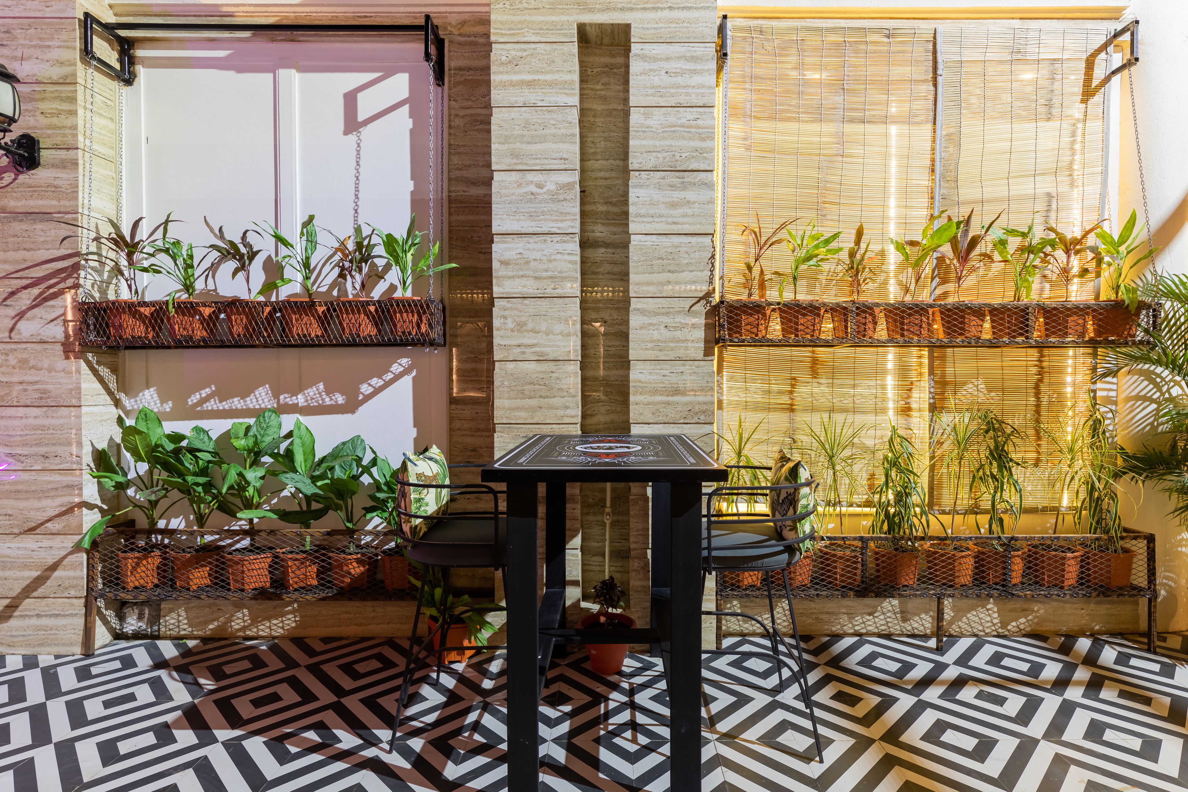 Un interior moderno con suelo de estampado geométrico, una mesa central y dos sillas metálicas. Hay plantas en macetas de alambre en las paredes.