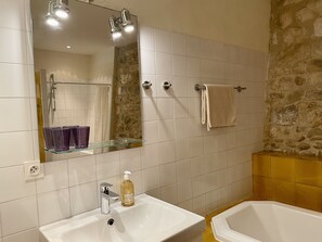 Ein Badezimmer mit einem weißen Waschbecken, einer Glasablage mit violetten Gläsern, einem Spiegel, einer Dusche mit Steinwand und einer Badewanne.