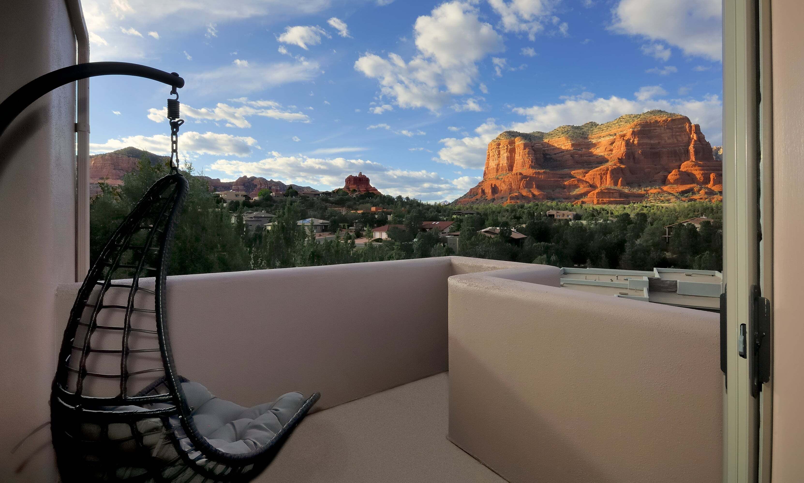 A balcony with a hanging chair, a view of red rock formations, and a residential area in the background.