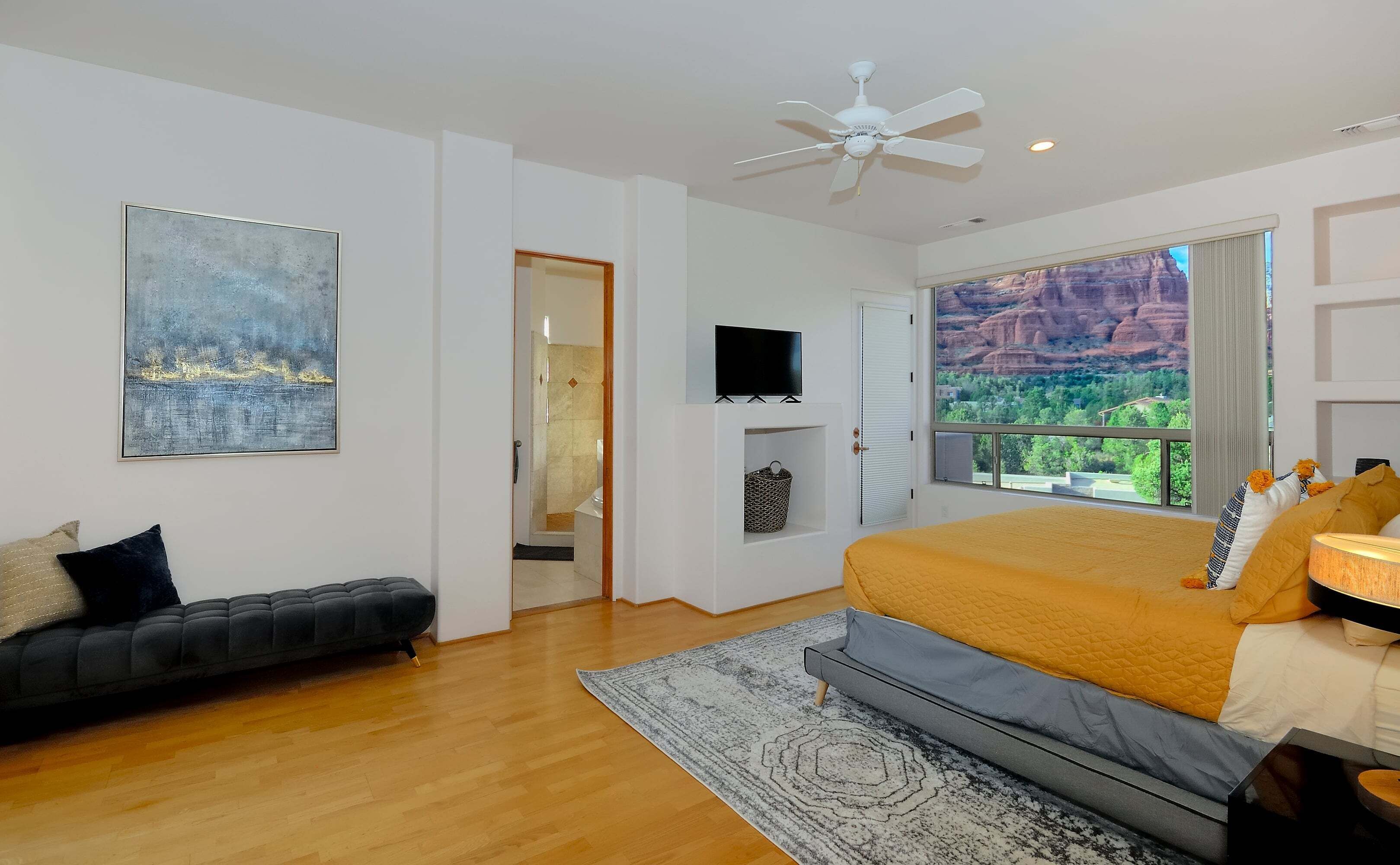 Quarto com cama, sofá, televisão, ventilador de teto e uma grande janela com vista para uma paisagem de montanhas.