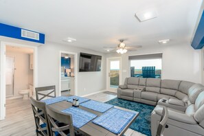 A modern living room with a beige sectional sofa, a dining table with blue placemats, and a ceiling fan. - Beach House II 103 - Picturesque;Beachfront Amenities;Sundeck;Huge pool (South Padre Island)