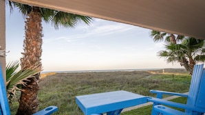 Ein überdachter Außenbereich mit blauen Stühlen und einem Tisch, mit Blick auf einen Strand und eine Rasenfläche.