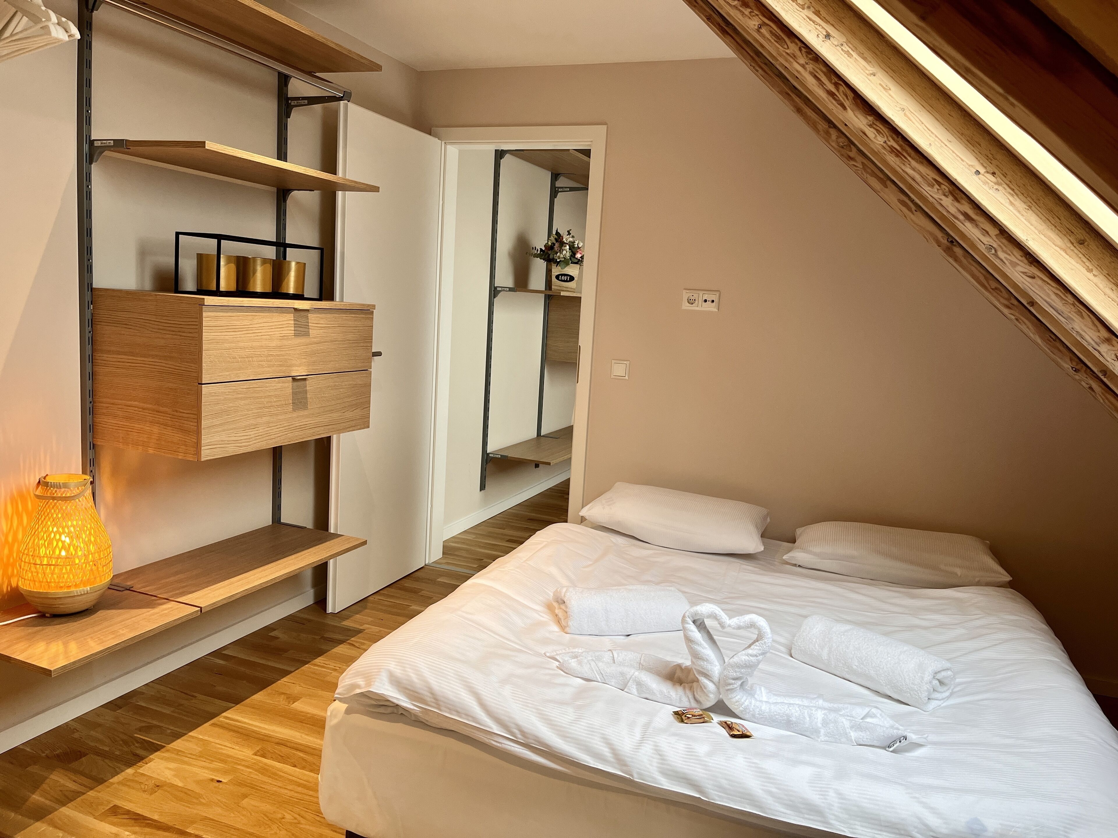 A bedroom with a bed, a wooden dresser, a hanging shelf, and a bedside table with a lamp.
